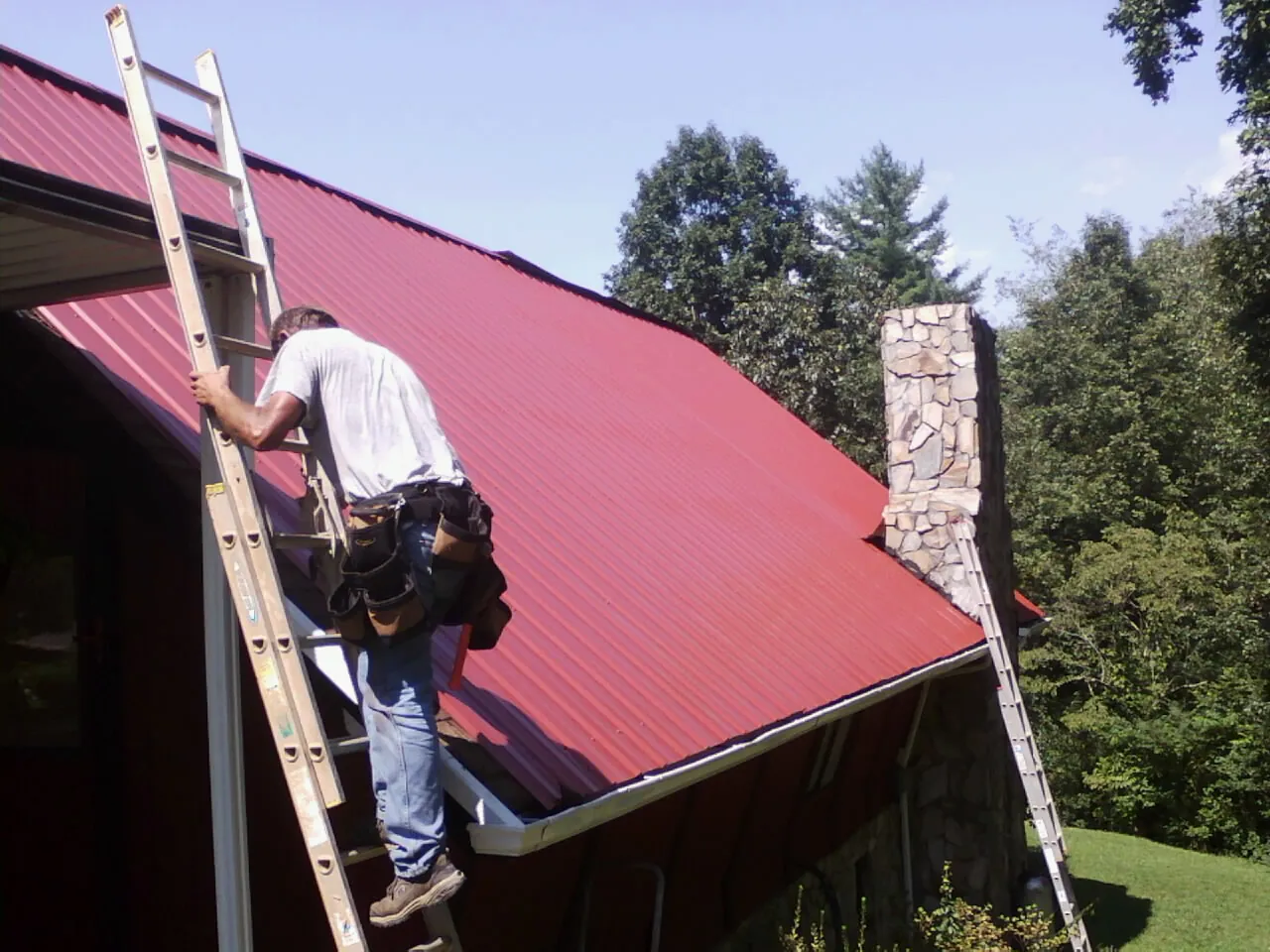 Roofer on a ladder beside a red metal roof and stone chimney — Soffit & Fascia Repair by Roofing Brothers in Loveland